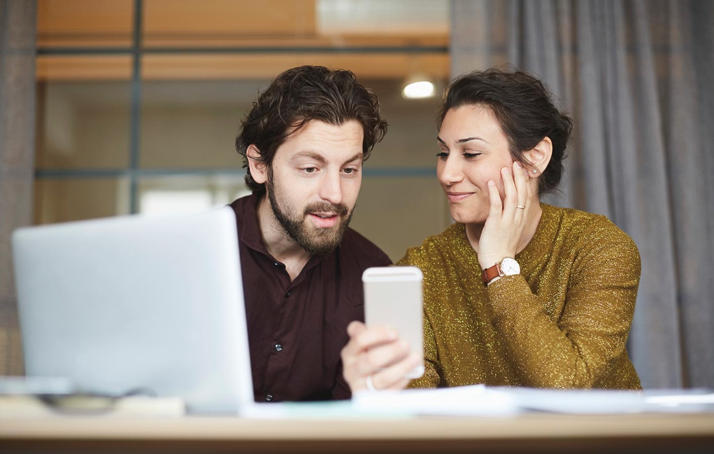 Mann und Frau sitzen zusammen an einem Tisch und schauen auf ein Telefon mit einem Notebook davor.